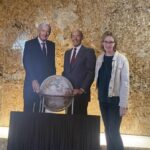 Kristin DuMez in white jacket beside two cutouts of Rich DeVos and Jay VanAndel who have their hands on a globe. 